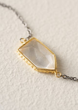 A close-up of a gold-toned pendant necklace with a faceted, translucent pale stone set on a textured cream background.