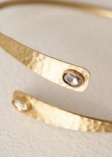 A hammered gold open cuff bangle bracelet with a small clear gemstone accent on each end, displayed on a textured off-white background.