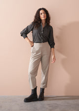 Woman wearing a check shirt, beige pants, and black boots against a beige wall.