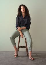 Woman sitting on a stool wearing a light cotton navy blouse with ruched cuffs, paired with green trousers and sandals