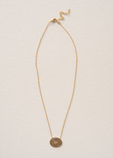 A delicate gold chain necklace with a small circular spiral pendant, displayed against a textured off-white background.