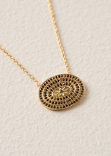 A gold oval pendant necklace featuring concentric rings of small black stones and a central floral motif, displayed on a textured off-white background.