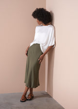 Woman wearing a white satin top and green skirt against a beige wall.