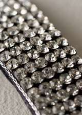 A close-up of a black fabric trim embellished with rows of faceted silver rhinestones, showing the reflective crystal details and the stitched edge.