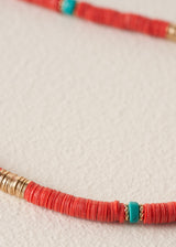 A close-up of a beaded necklace featuring textured coral-red cylindrical beads, accented with small turquoise blue beads and gold-toned spacer rings, laid on a light beige textured surface.