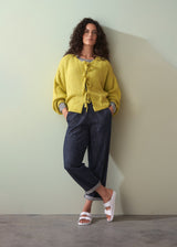woman leaning against a green background, wearing an acid yellow knit cardigan with bow tie front, paired with navy trousers and white sandals