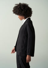 Woman wearing a black relaxed blazer, white shirt and black trousers against a light gray background