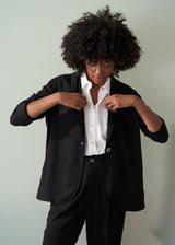 Woman wearing a black relaxed blazer, white shirt and black trousers against a light gray background