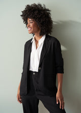 Woman wearing a black relaxed blazer, white shirt and black trousers against a light gray background