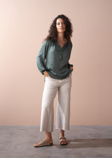 Woman wearing a green blouse and off white wide leg trousers against a beige wall