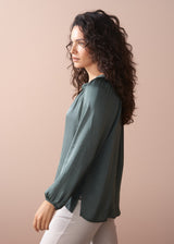 Woman wearing a green blouse and off white wide leg trousers against a beige wall