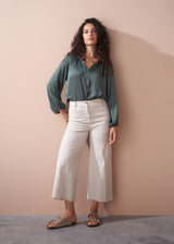 Woman wearing a green blouse and off white wide leg trousers against a beige wall