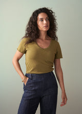 Woman wearing an olive green v-neck t-shirt with blue jeans and gold bangle, posing with her hand tucked in a pocket