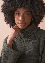Woman wearing a green chunky knit toll neck sweater and oatmeal cord trousers against a beige background
