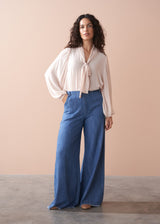 Woman wearing a light pink pussybow blouse and blue wide leg jeans against a beige background