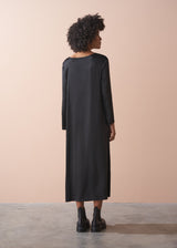 Woman wearing a round neck black satin dress and black leather boots, standing against a beige background