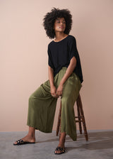 Woman wearing a black top and green wide leg satin trousers sitting on a wooden stool against a beige background