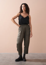 Woman wearing a black camisole top and olive green satin trousers against a beige background