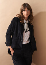 Woman wearing a black corduroy blazer, white shirt, and patterned tie against a beige background