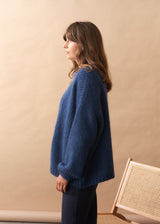Woman wearing a chunky knit blue sweater standing against a beige wall with a wicker chair.
