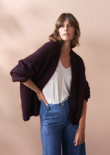 Woman wearing a dark purple chunky knit cardigan over a white top and blue jeans on a beige background.