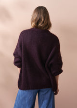 Person wearing a dark purple chunky knit sweater and blue jeans against a beige background.