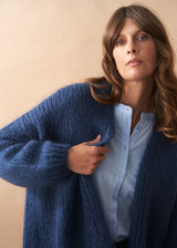 Woman wearing a chunky knit blue cardigan over a white shirt against a beige background