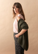 Woman wearing a green cardigan over a pink top and patterned pants against a beige background