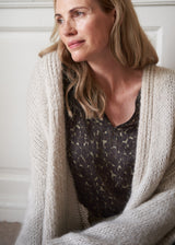 A woman wearing a chunky cream-colored knit cardigan over a dark patterned top, captured in a soft, warm indoor setting.