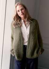 A woman wearing a chunky khaki green cable-knit cardigan over a white button-down shirt and dark blue jeans, standing in a doorway.