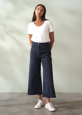 Woman wearing a white t-shirt and dark blue wide leg cropped trousers standing against a plain background