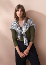 Woman wearing a green cashmere blend top and black pants with a striped jumper against a beige background