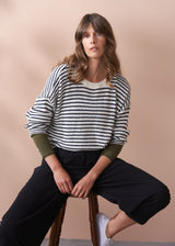 Woman wearing a striped sweater and black trousers sitting on a stool against a beige background