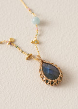 A gold-toned beaded necklace featuring a large teardrop-shaped labradorite pendant with blue-green iridescence, accented with small colorful beads on a textured cream background.