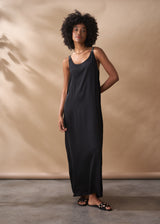 A woman in a scoop-neck black satin maxi dress stands against a beige background.