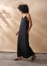 A woman in a scoop-neck black satin maxi dress stands facing a beige background.