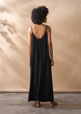 A woman in a scoop-back black satin maxi dress stands facing a beige background.