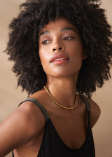 Close-up of a woman in a scoop-neck black satin maxi dress stands with gold necklace, against a beige background.