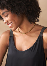 Close up of a woman in a scoop-neck black satin maxi dress with gold necklace, against a beige background.