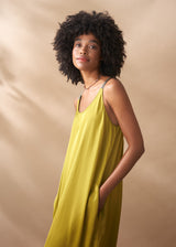 A woman in a scoop-neck yellow satin maxi dress stands with her hands in her pockets against a beige background.
