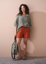Woman wearing a green sweater and orange shorts holding a green bag against a beige wall.