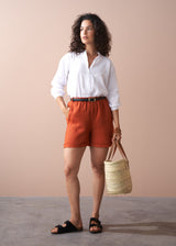 Woman wearing a white shirt and orange shorts, holding a straw bag against a beige background