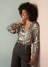Woman wearing a patterned blouse and dark pants against a light wall.