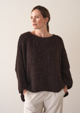 Woman wearing a dark brown boucle sweater and cream trousers against a plain background