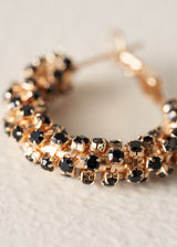 A close-up of a gold-toned hoop earring embellished with multiple round black faceted gemstones, set against a soft, light beige textured background.