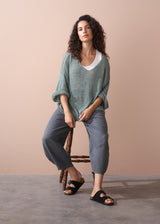 Woman sitting on a wooden stool wearing a green sweater and checkered linen trousers against a beige background
