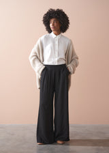 Person wearing a white shirt and beige sweater with black wide-leg trousers against a plain background