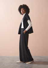 Person wearing a black boucle gilet jacket over a cream poloneck and black trousers against a beige background