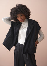 Person wearing a black boucle gilet jacket over a cream poloneck and black trousers against a beige background