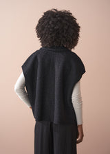 Person wearing a black boucle gilet jacket over a cream poloneck and black trousers against a beige background